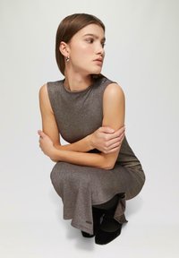 Metaliczna brązowa sukienka o dopasowanej sylwetce i rozkloszowanym dolnym brzegu. Modelka ma na sobie czarne baleriny oraz proste srebrne kolczyki.