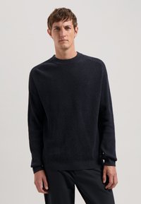 Pulover din lână în nuanță închisă de navy, cu guler rotund, textură bordeiată, mâneci lungi și mansete strâmte, asortat cu pantaloni negri. Design minimalist.