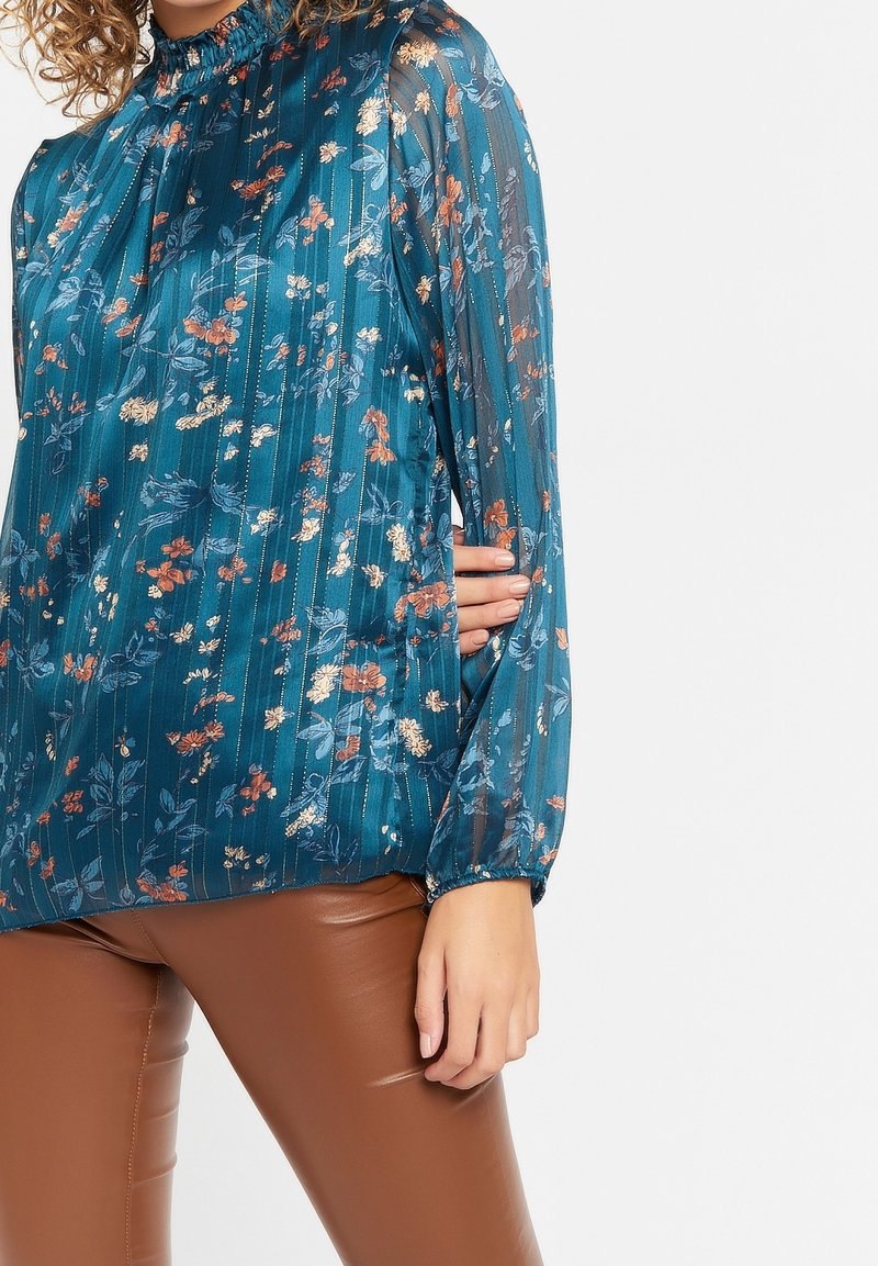 Tursblå blus med blommönster och plisserad design, hög hals och långa ärmar; har gyllene detaljer och en genomskinlig textur.