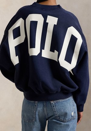 Persoon draagt een marineblauwe sweatshirt met grote witte letters "POLO" op de rug en lichtblauwe spijkerbroek tegen een effen achtergrond.