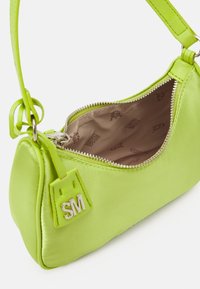 Sac à main en satin vert lime vif avec une forme arrondie, fermeture éclair et petit patch logo. Il dispose d'une courte bandoulière et d'un doublure intérieure douce.