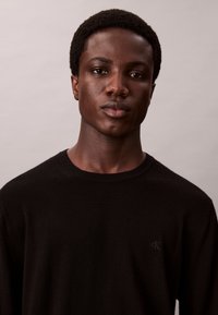 Jeune homme noir aux cheveux courts et bouclés, portant un pull noir à col rond, faisant face à l'objectif avec une expression neutre sur un fond uni.