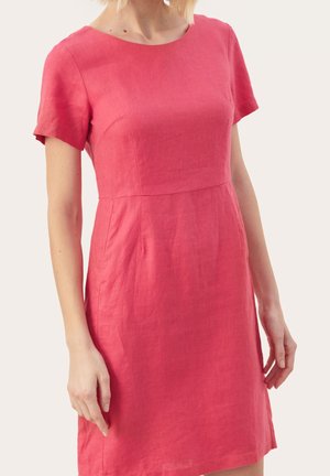 Frau trägt ein einfarbiges, korallenrosa Kleid mit kurzen Ärmeln und knielangem Schnitt sowie Rundhalsausschnitt, steht vor neutralem Hintergrund.