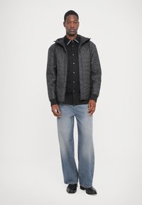 Schwarze gesteppte Jacke mit Kapuze, langen Ärmeln und gerippten Bündchen, getragen über einem schwarzen Hemd mit Knöpfen und hellblauen weit geschnittenen Jeans.