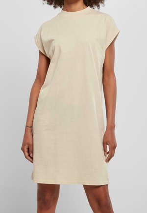 Vrouw draagt een knielange, beige mouwloze T-shirtjurk met opgerolde mouwen, staand voor een effen lichtgrijze achtergrond.