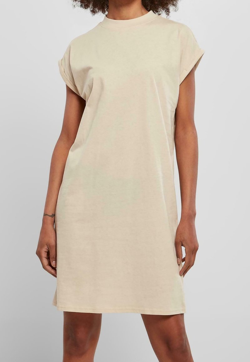 Femme portant une robe T-shirt beige sans manches, longueur genoux, avec manches retroussées, debout devant un fond uni gris clair.