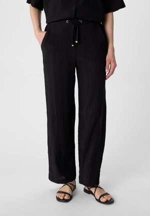 Black, wide-leg pants with a textured fabric, drawstring waist, and side pockets, paired with black strappy sandals.