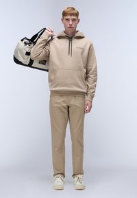 Sudadera beige con capucha y bolsillo frontal, combinada con pantalones beige y zapatillas blancas. Lleva una bolsa de viaje negra y beige con un parche de bandera.