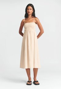 Lys beiges kjole med en tætsiddende, smocket overdels og tynde stropper, der falder til midt på læggen. Stoffet ser glat og let ud.