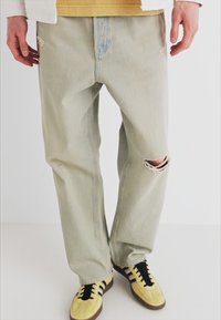 Lys blå, slitt denim jeans med løs passform, med detaljer som har rifter ved knærne. Brukt sammen med gule sneakers og en gul skjorte.