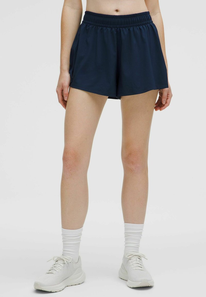 Marinblå träningsshorts med elastisk midja, med en jämn textur och avslappnad passform, tillsammans med vita träningsskor.