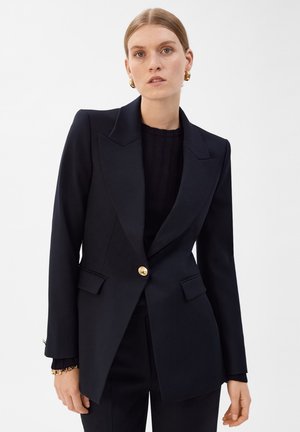 Femme aux cheveux blonds porte un blazer noir ajusté avec un bouton doré et un pantalon noir assorti sur fond uni.