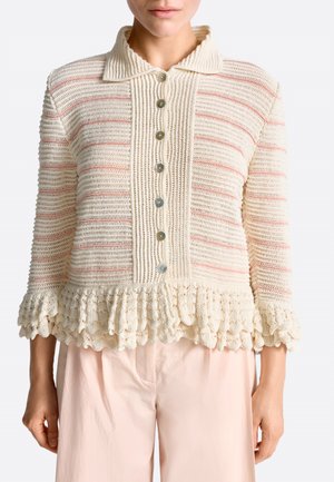 Woman wearing a cream crochet cardigan with buttons, ruffled hem and cuffs, paired with pale pink pants, shown from neck to hips.