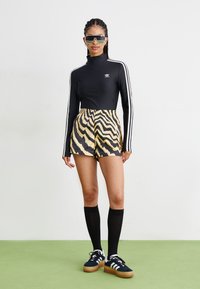 Zwarte lange mouwen top met witte strepen, tijgergestreepte shorts in geel en zwart, zwarte kniehoogsokken, en zwarte sneakers met witte accenten.