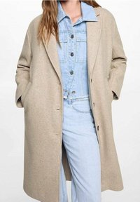 Béžový vlněný kabát s volným střihem, vybavený klopovým límcem, knoflíky vpředu a bočními kapsami, nošený přes světle modrý denimový outfit.