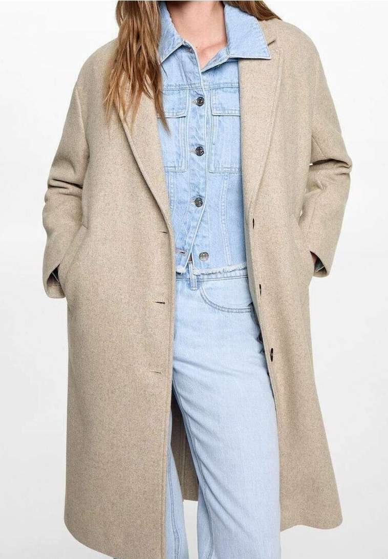 Béžový vlněný kabát s volným střihem, vybavený klopovým límcem, knoflíky vpředu a bočními kapsami, nošený přes světle modrý denimový outfit.