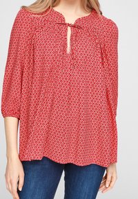 Blouse florale rouge avec un décolleté en trou de serrure, des détails froncés et des manches trois-quarts. Présente un motif circulaire blanc sur l'ensemble.