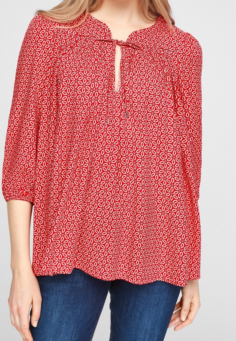 Blouse florale rouge avec un décolleté en trou de serrure, des détails froncés et des manches trois-quarts. Présente un motif circulaire blanc sur l'ensemble.
