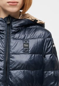 Γυναίκα που φοράει μπλε σκούρο γυαλιστερό μπουφάν puffer με μπεζ κουκούλα και μαύρο φερμουάρ, με ετικέτες «Blauer U.S.A.» στο στήθος και στο μανίκι.