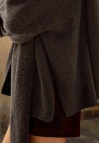 Grauer, weicher Strickpullover mit lockerer Passform, der ein drapiertes Design und lange Ärmel aufweist. Kombiniert mit dunkelbraunen Cordshorts.
