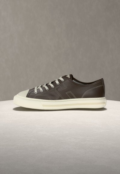 Zapatilla baja de color marrón oscuro con cordones blancos, puntera de goma blanca y suela blanca gruesa sobre un fondo neutro.