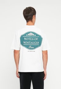 White cotton t-shirt featuring a large teal graphic on the back with the text "NOTES OF NOSTALGIA" and "DON'T WASTE CULTURE." Short sleeves.