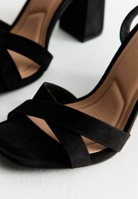 Sandalias de tacón alto de ante negro con punta cuadrada y amplias correas entrelazadas. Cuentan con un tacón grueso y una plantilla suave de color beige.