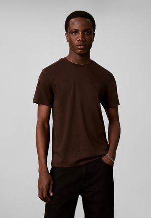 Joven con una camiseta lisa de manga corta de color marrón oscuro y pantalones negros, de pie con una mano en el bolsillo sobre un fondo gris.