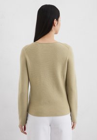 Long-sleeve sweater in light olive green with a ribbed texture, round neckline, and curved hem, paired with white pants.