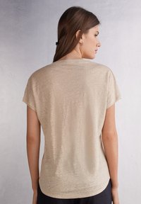Beige top s krátkým rukávem vyrobený z měkké texturované látky a s kulatým výstřihem. Zadní strana má hladký povrch a uvolněný tvar.