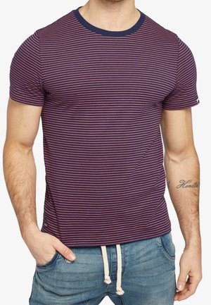 Mand iført en tætsiddende kortærmet t-shirt med lilla og pink striber, navy krave og lyseblå jeans med snor.