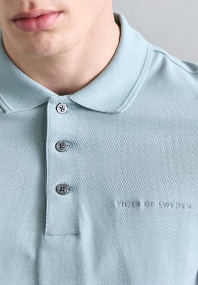 Svetlo modra polo majica iz teksturiranega blaga, z ovratnikom, tremi gumbi in odtisnjeno logotipom: "TIGER OF SWEDEN."