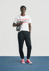 Λευκό βαμβακερό μπλουζάκι με κόκκινες και μπλε ρίγες, μαύρο παντελόνι jogging και κόκκινα αθλητικά παπούτσια. Το μοντέλο κρατάει μια μικρή μαύρη τσάντα χιαστί.