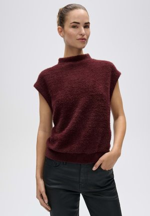 Donna con i capelli raccolti indossa un top senza maniche peloso color bordeaux e pantaloni neri, in piedi con una mano in tasca su uno sfondo semplice.