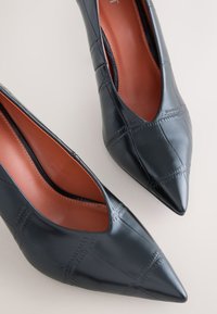 Paire de chaussures pour femmes en cuir noir à bout pointu, avec motif cousu et semelles intérieures en cuir marron sur fond clair.