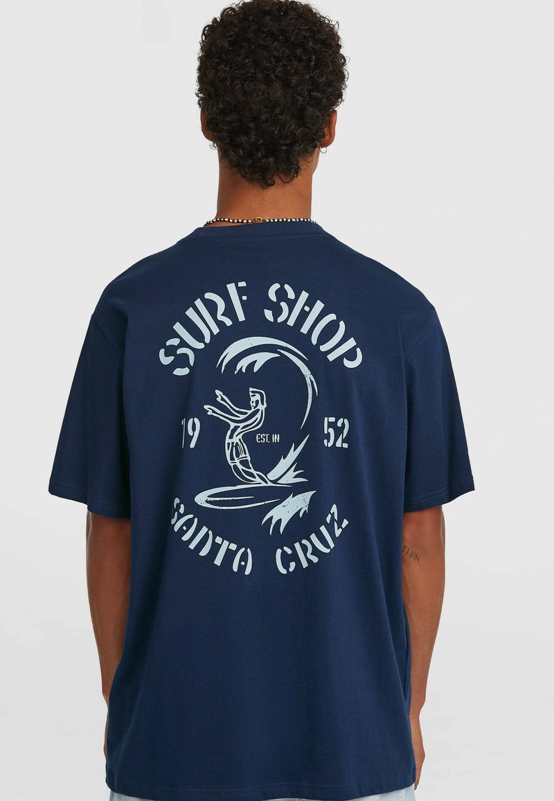 Μπλε βαμβακερό T-shirt με γραφική σχεδίαση στην πλάτη που περιλαμβάνει το κείμενο "SURF SHOP SANTA CRUZ" και ένα θέμα σερφ, άνετη εφαρμογή, κοντά μανίκια.