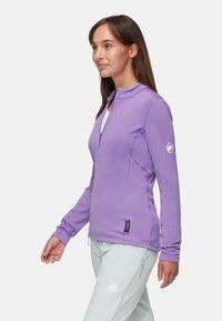 Ljust lila kvartssämpelpullover med långa ärmar, tillverkad av ett mjukt och stretchtejligt tyg. Logotypdetalj på ärmen, bärs med ljusgrå byxor.