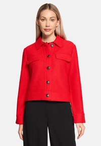 Rode, korte jacket gemaakt van textiel. Voorzien van een kraag, sluiting met knopen aan de voorkant en twee borstzakken met zwarte knopen.