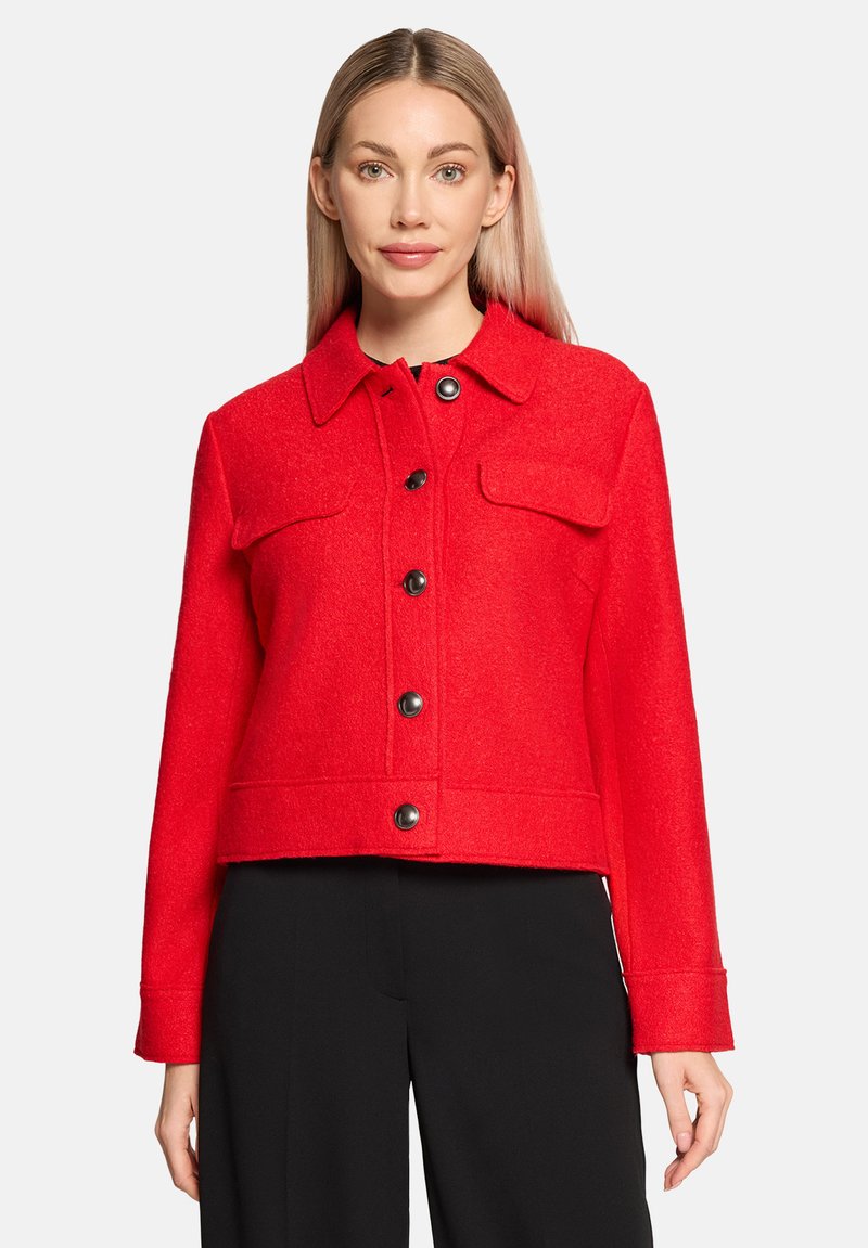 Rode, korte jacket gemaakt van textiel. Voorzien van een kraag, sluiting met knopen aan de voorkant en twee borstzakken met zwarte knopen.