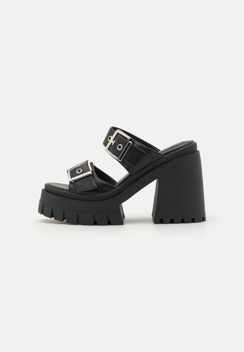 Sandales à plateforme noires avec une semelle épaisse et crantée et un talon épais. Elles sont dotées de deux larges sangles avec des boucles en argent et des accents circulaires.