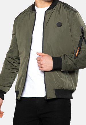 Homme portant une veste aviateur vert olive avec des poignets noirs, une fermeture éclair et un petit patch rond sur la poitrine, par-dessus une chemise blanche et un pantalon noir.