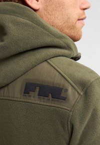 Olijfgroen fleecjas met een gestructureerde capuchon, voorzien van een zwart logo-label en contrasterende gladde schouderpanelen. Accenten in de stiksels zijn zichtbaar.
