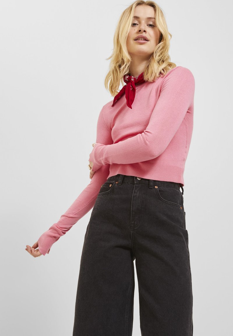 Maglione aderente rosa a maniche lunghe con polsini a coste, abbinato a jeans neri a vita alta e a gamba larga. Un bandana rossa è annodata al collo.