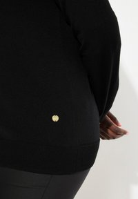 Svart stickad tröja med en strukturerad yta. Har en guldig logotypdetalj på den nedre sidan. Fitted silhuett med ribbad fåll.