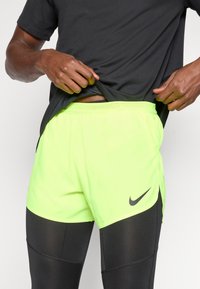 Uomo che solleva la maglietta nera per rivelare dei pantaloni corti Nike giallo neon indossati sopra dei leggings sportivi neri.