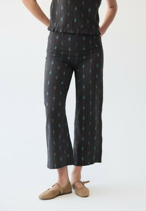 Person trägt schwarze Hose und passendes Oberteil mit vertikalem türkis- und grau-floralem Muster, kombiniert mit hellbraunen Mary Jane Schuhen.