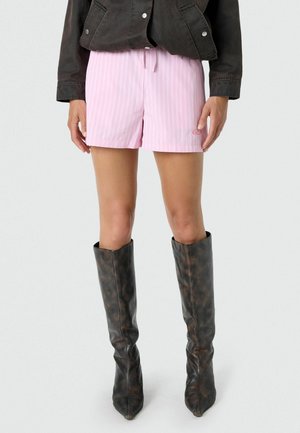 Persona con pantalones cortos de rayas rosa claro, chaqueta negra y botas altas de cuero marrón oscuro, de pie contra un fondo liso.