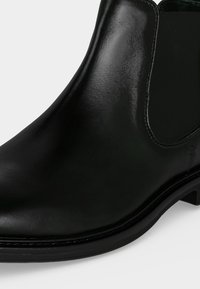 Gros plan d'une botte Chelsea en cuir noir avec un panneau latéral élastique et un petit talon sur fond blanc.