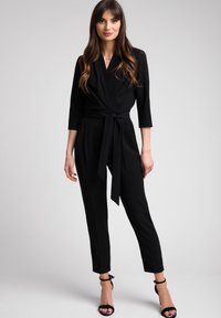 Czarny kombinezon z wiązaniem, długimi rękawami i paskiem w talii. Dopasowany krój z zwężającymi się nogawkami, wykonany z gładkiego materiału bez widocznych wzorów.