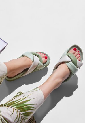 Green and white slide sandals with braided straps, soft textured footbed, and red toenail polish on exposed feet, paired with printed pants.